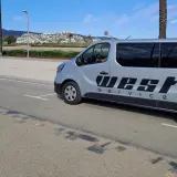 Srebrny bus z napisem "WEST SERVICE" gotowy do międzynarodowego przewozu osób. W tle palmy i zabudowania na wzgórzu.