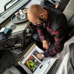 Kierowca ciężarówki przygotowuje się do dobowego odpoczynku, sięgając po napoje z lodówki. Na desce rozdzielczej tablet i nawigacja.