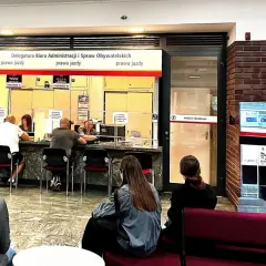 Ludzie czekają w kolejce do okienek w urzędzie, gdzie można załatwić sprawy związane z prawem jazdy. Na tablicy informacyjnej widnieje nr PKK.