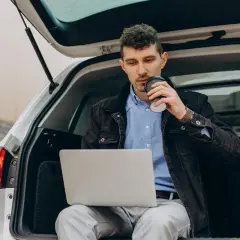 Mężczyzna siedzi w bagażniku samochodu z laptopem i kawą. Wygląda na to, że przekroczenie czasu pracy o 2 godziny nie jest dla niego problemem.