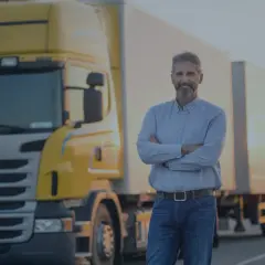 Uśmiechnięty kierowca ciężarówki złożył ręce na piersi. Obok stoi żółta ciężarówka. OCP co to? To jego praca.