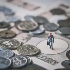 Mała figurka biznesmena stoi wśród japońskich monet i banknotów, symbolizując znaczenie **diet krajowych** w jego podróży.