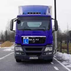 Niebieska ciężarówka MAN z literą "L" na grillu, gotowa do egzaminu C+E. Na placu manewrowym śnieg i pachołki.