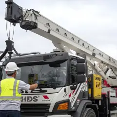 Pracownik w kamizelce odblaskowej i kasku sprawdza HDS. Na placu budowy stoi podnośnik koszowy.