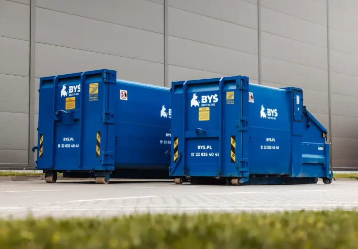 Dwa niebieskie kontenery BYS Recycling stoją przed budynkiem. Oferują tani transport odpadów.