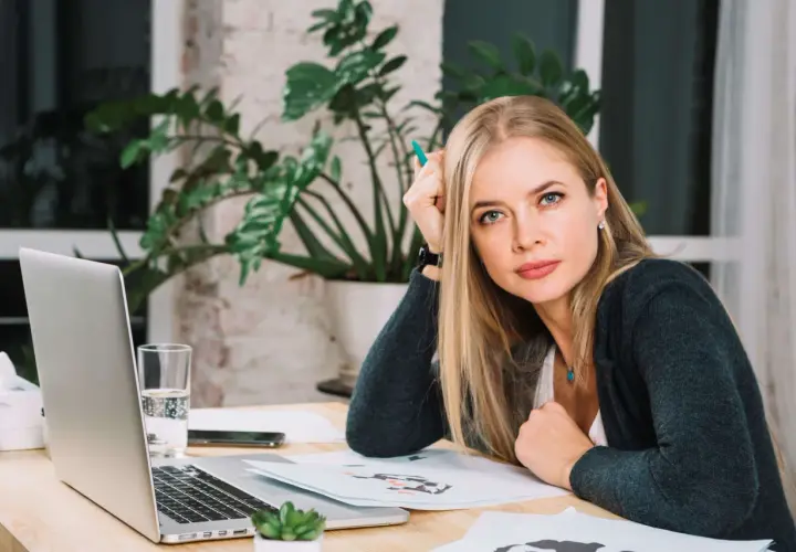 Blondynka z zielonym długopisem w dłoni, zastanawia się nad różnymi rodzajami odpoczynku, siedząc przy biurku z laptopem.