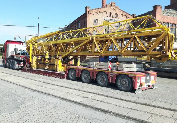 Ciężarówka z przyczepą transportuje żółty element dźwigu. Wymaga to zezwolenia na transport ponadgabarytowy.