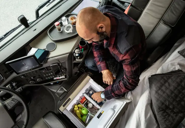 Kierowca ciężarówki przygotowuje się do dobowego odpoczynku, sięgając po napoje z lodówki. Na desce rozdzielczej tablet i nawigacja.