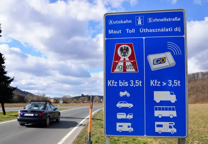 Znak informuje o opłatach za autostrady w Austrii. Widoczny jest samochód na drodze i niebieski znak z symbolami pojazdów.