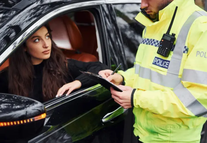 Policjant w kamizelce odblaskowej spisuje mandat za wagę. Kobieta w samochodzie patrzy z niepokojem.