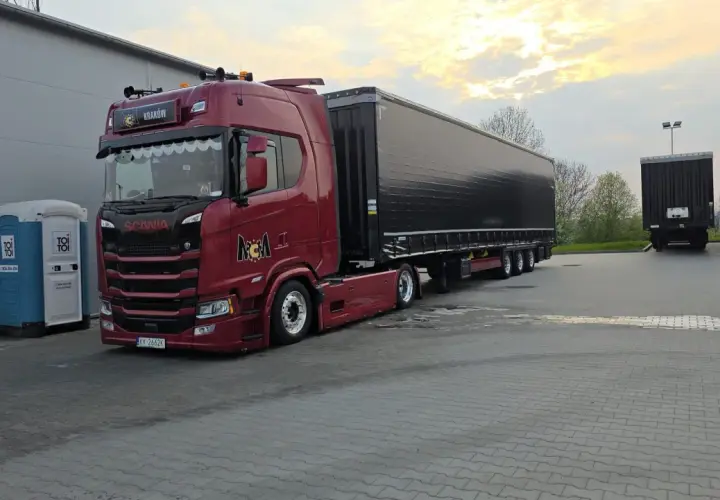 Czerwona Scania z naczepą gotowa do przewozu drobnicowego. Obok stoi niebieska toaleta przenośna.