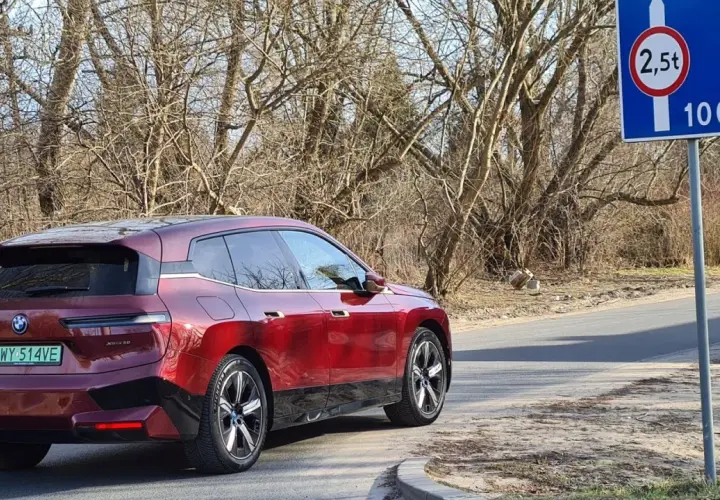 Czerwony BMW iX jedzie drogą, zbliżając się do znaku informującego o zakazie tonażowym 2,5 tony na dystansie 100m.