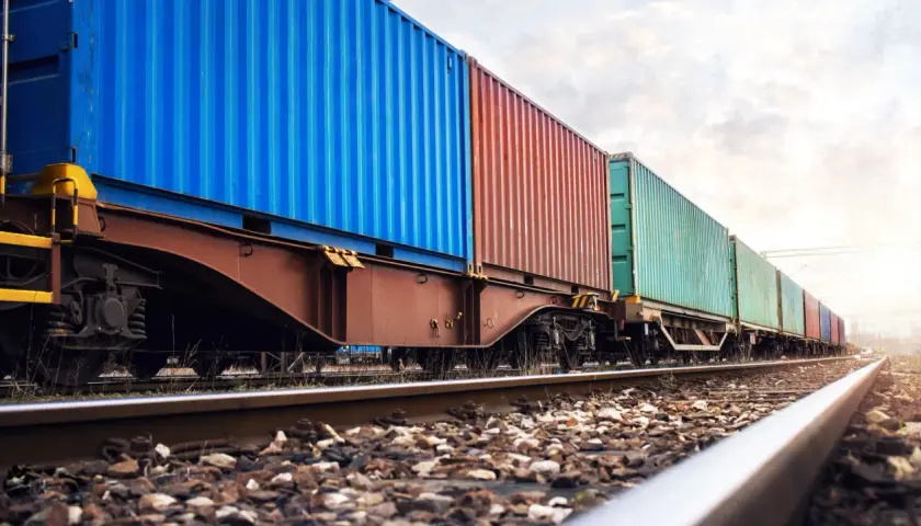 Długi pociąg towarowy z kolorowymi kontenerami, symbolizujący globalne konwencje transportowe, jedzie po torach w słoneczny dzień.