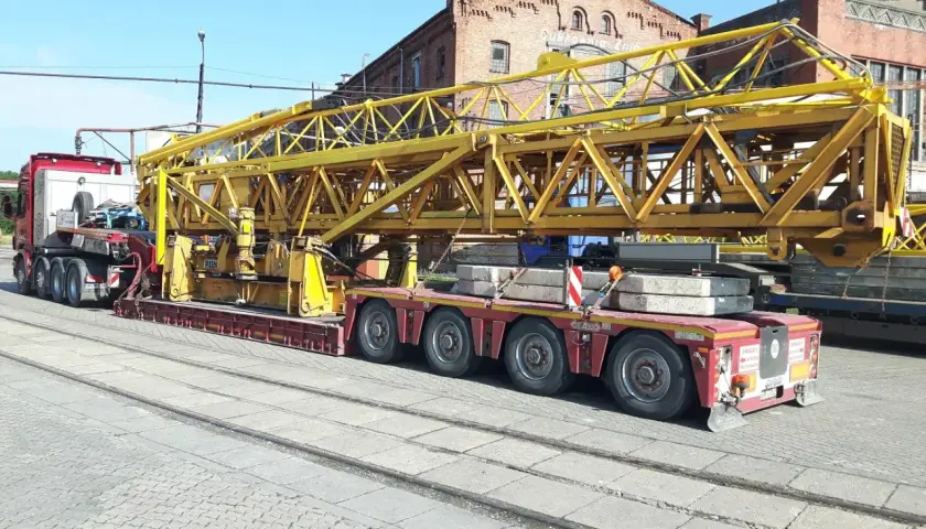 Ciężarówka z przyczepą transportuje żółty element dźwigu. Wymaga to zezwolenia na transport ponadgabarytowy.