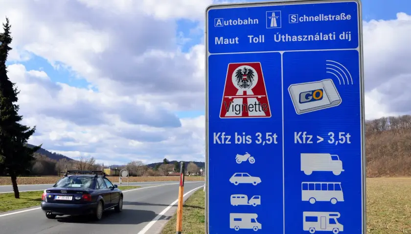 Znak informuje o opłatach za autostrady w Austrii. Widoczny jest samochód na drodze i niebieski znak z symbolami pojazdów.