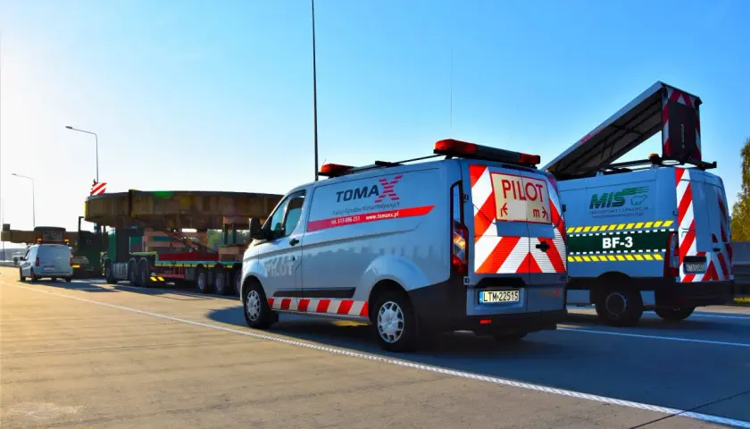 Pojazd ponadgabarytowy transportowany na platformie, eskortowany przez dwa samochody pilotujące z napisem "PILOT".