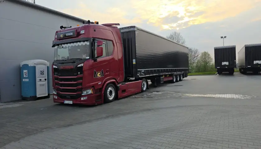 Czerwona Scania z naczepą gotowa do przewozu drobnicowego. Obok stoi niebieska toaleta przenośna.