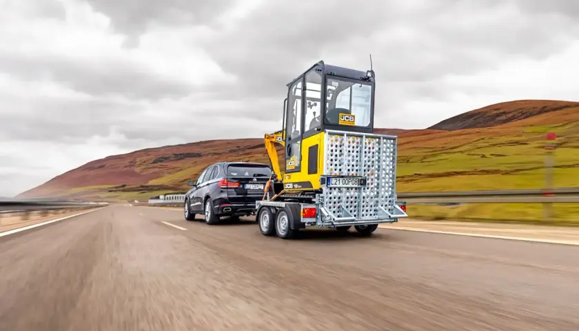 Czarny SUV ciągnie na lawecie minikoparkę JCB. Czy na lawetę trzeba mieć uprawnienia?