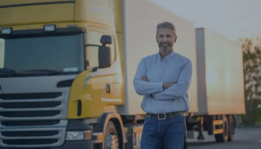 Uśmiechnięty kierowca ciężarówki złożył ręce na piersi. Obok stoi żółta ciężarówka. OCP co to? To jego praca.