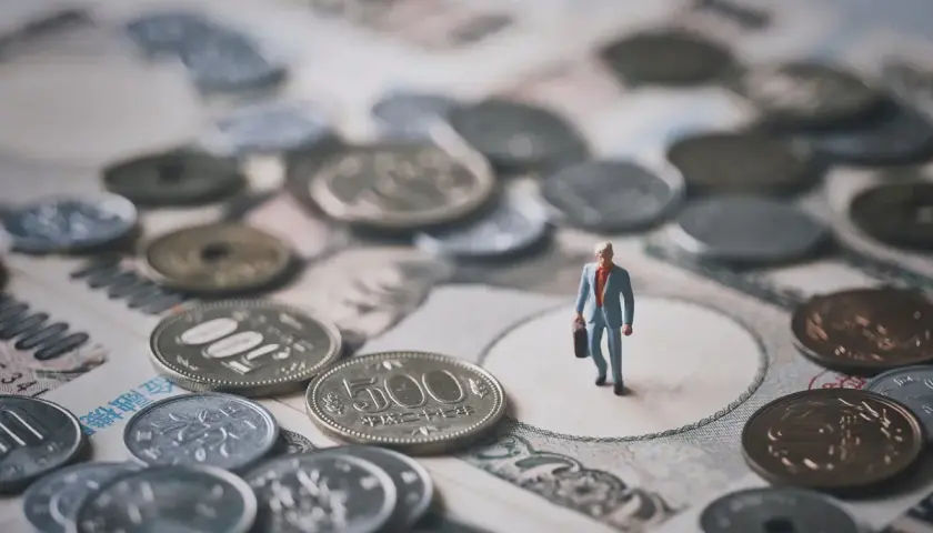 Mała figurka biznesmena stoi wśród japońskich monet i banknotów, symbolizując znaczenie **diet krajowych** w jego podróży.