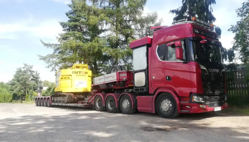 Czerwony Scania z naczepą transportuje żółtą koparkę. To typowa przesyłka dużych gabarytów, gotowa do drogi.