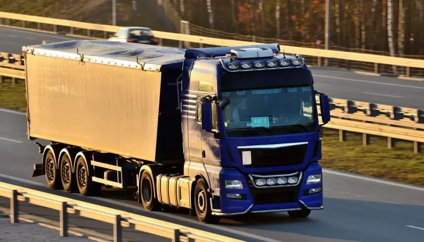 Niebieska ciężarówka z naczepą jedzie autostradą. Kierowca z kodem 95 prawo jazdy wiezie towar.