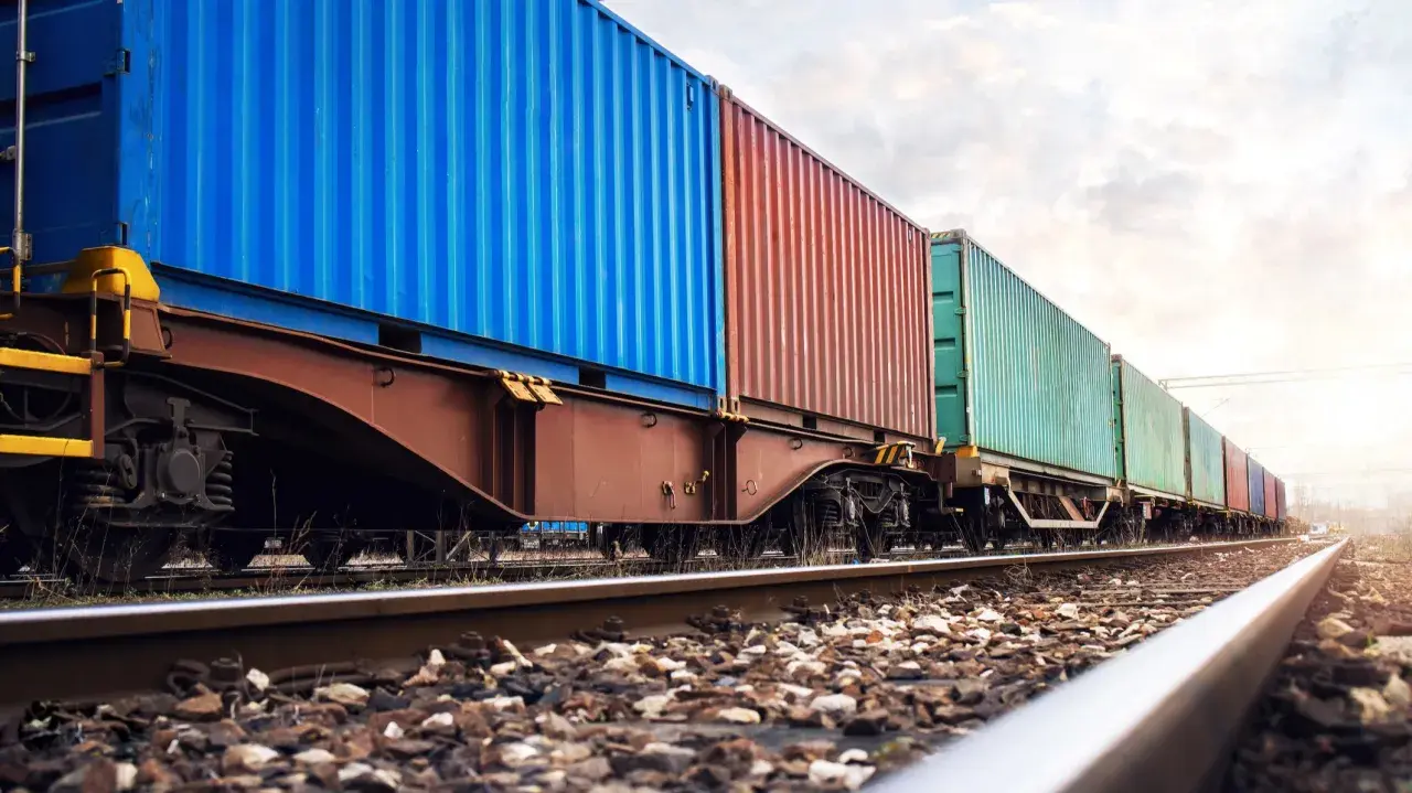 Długi pociąg towarowy z kolorowymi kontenerami, symbolizujący globalne konwencje transportowe, jedzie po torach w słoneczny dzień.