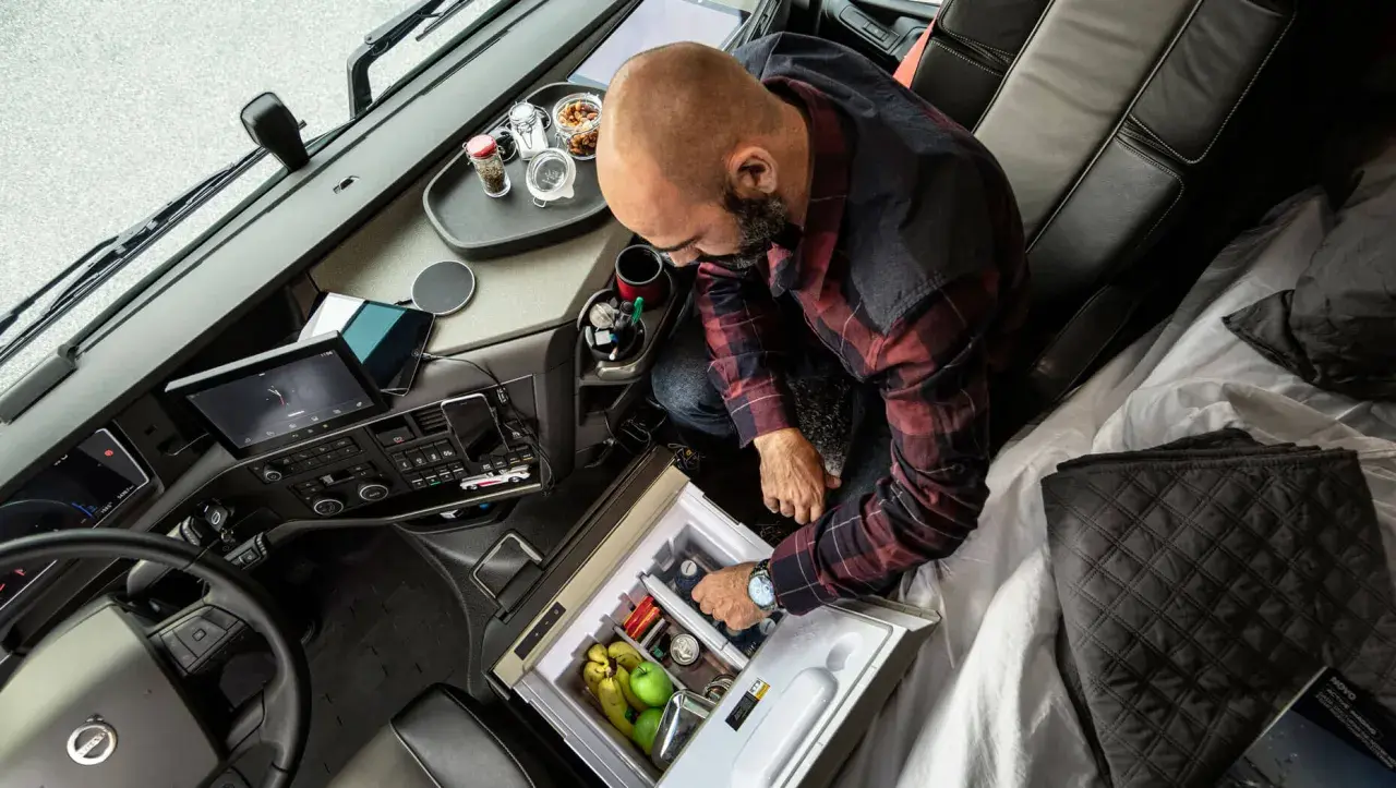 Kierowca ciężarówki przygotowuje się do dobowego odpoczynku, sięgając po napoje z lodówki. Na desce rozdzielczej tablet i nawigacja.