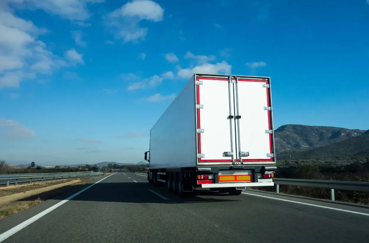 Biała naczepa ciężarówki na autostradzie, przewożąca fracht. Co to jest fracht? To towar w transporcie.