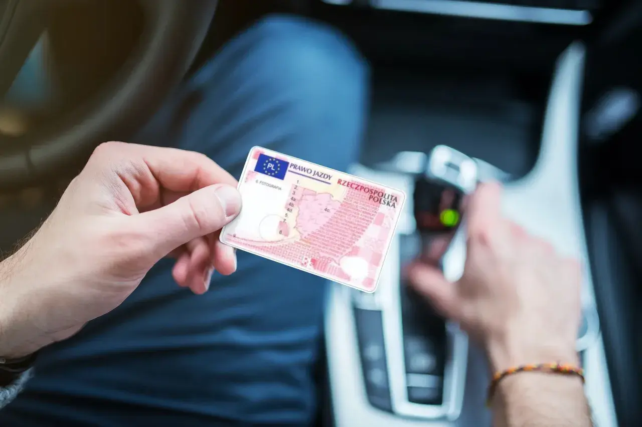 Osoba trzyma polskie prawo jazdy. W tle widać wnętrze samochodu, co sugeruje, że może to być dokument potrzebny do prowadzenia pojazdu, gdy posiada się prawo jazdy spoza Unii Europejskiej.
