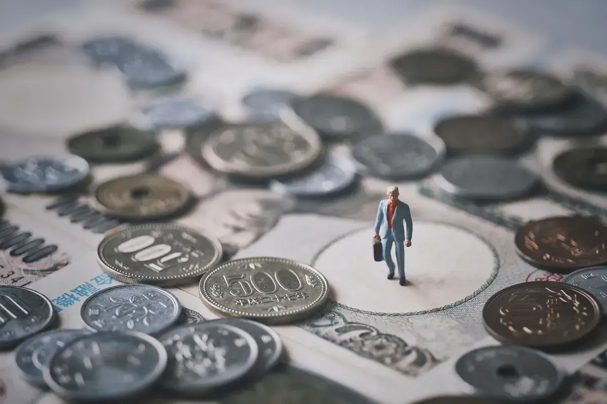 Mała figurka biznesmena stoi wśród japońskich monet i banknotów, symbolizując znaczenie **diet krajowych** w jego podróży.