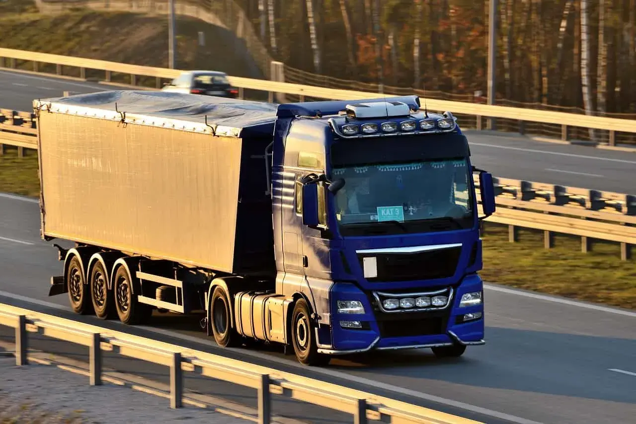 Niebieska ciężarówka z naczepą jedzie autostradą. Kierowca z kodem 95 prawo jazdy wiezie towar.