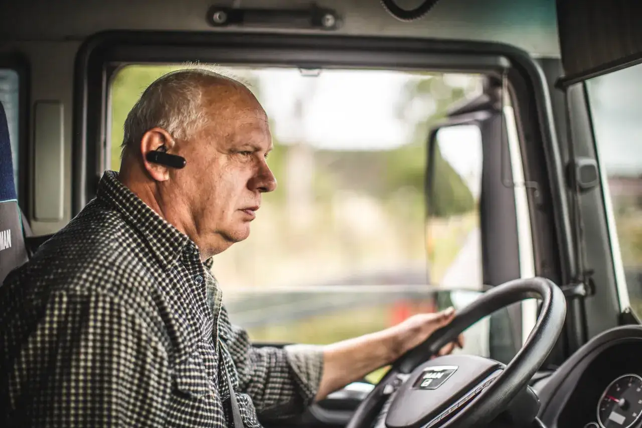 Starszy mężczyzna, jeden z wielu kierowców z Ukrainy, prowadzi ciężarówkę, słuchając czegoś przez słuchawkę Bluetooth.