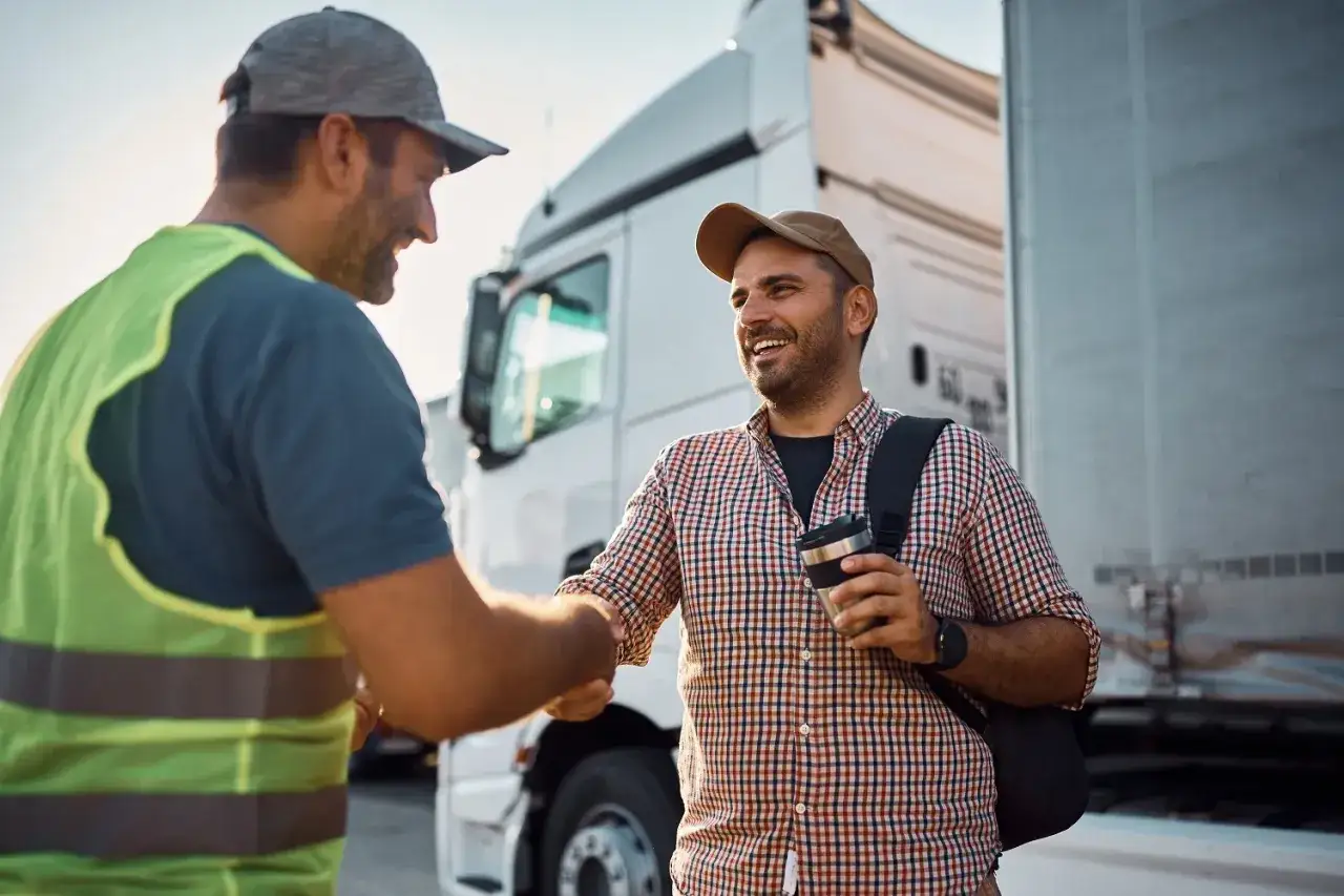 Dw&oacute;ch kierowc&oacute;w ciężar&oacute;wek wita się na tle pojazd&oacute;w. Jeden z nich ma na sobie kamizelkę odblaskową. To obraz sukcesu szkoleń dla firm transportowych.