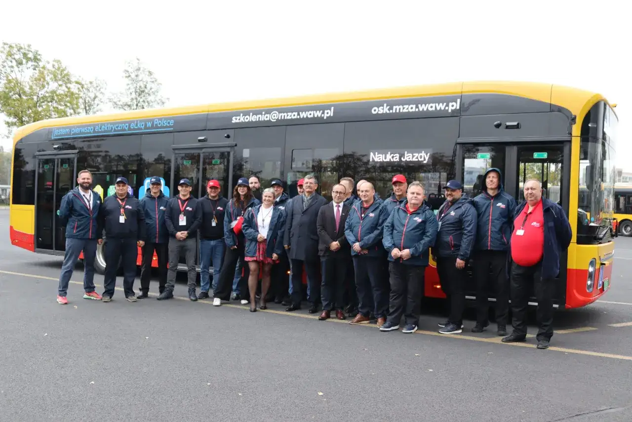 Grupa os&oacute;b pozuje przed nowym autobusem elektrycznym. Na boku pojazdu widnieje napis 