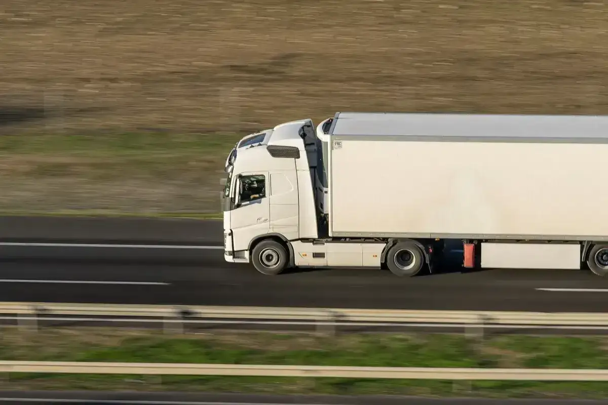 Biała ciężar&oacute;wka z naczepą jedzie autostradą. Usługa kierowania pojazdem powierzonym PKD w ruchu.